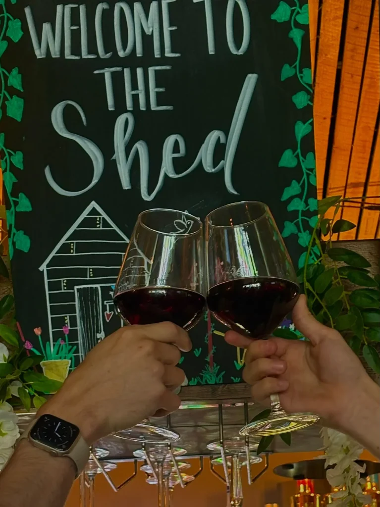 Two glasses of red wine raised in front of the Welcome to The Shed chalkboard